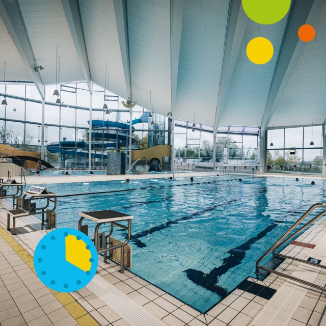 Blick auf das Sportbecken im Triamare mit blauem Wasser, markierten Schwimmbahnen, Startblöcken am Beckenrand und großen Glasfenstern. Im Hintergrund ist die blaue Wasserrutsche zu sehen. Unten links zeigt ein blaues Uhrsymbol mit gelbem Segment eine Aufe