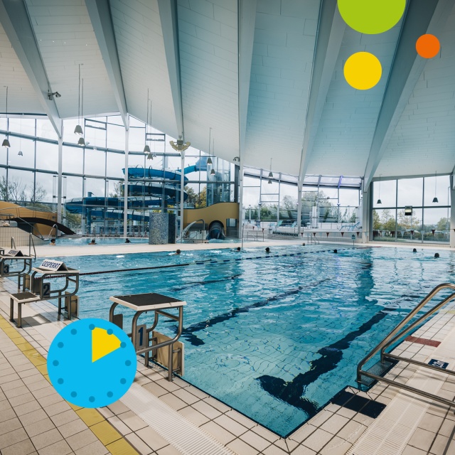 Blick auf das Sportbecken im Triamare mit blauem Wasser, markierten Schwimmbahnen, Startblöcken am Beckenrand und großen Glasfenstern. Im Hintergrund ist die blaue Wasserrutsche zu sehen. Unten links zeigt ein blaues Uhrsymbol mit gelbem Segment eine Aufe