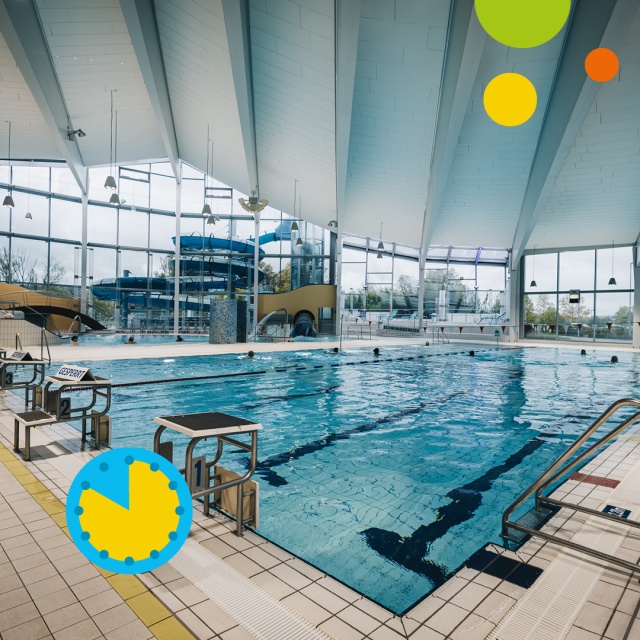 Blick auf das Sportbecken im Triamare mit blauem Wasser, markierten Schwimmbahnen, Startblöcken am Beckenrand und großen Glasfenstern. Im Hintergrund ist die blaue Wasserrutsche zu sehen. Unten links kennzeichnet ein blaues Uhrsymbol mit großer gelber Flä